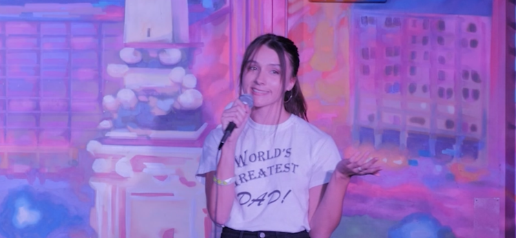Ariana performs standup comedy wearing a "World's Greatest Dad!" t-shirt. The stage background is a purple and pink mural of Indianapolis
